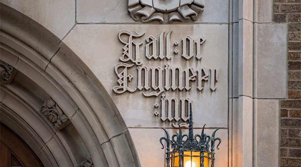 A decorative stone relief reading "Hall of Engineering" on the facade of Cushing Hall of Engineering at the University of Notre Dame. Below the relief, a detailed metal lantern emits a warm light, complementing the building's traditional architectural design.
