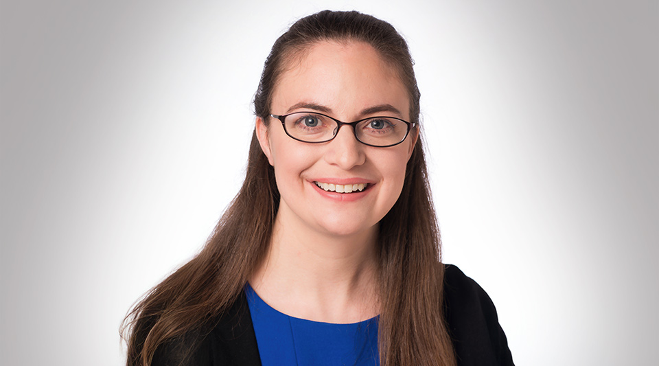 Portrait of Professor Ashley Thrall, smiling and wearing glasses, a blue top, and a black blazer, against a light gray background.