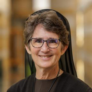 A professional portrait of Sr. Damien Marie Savino, FSE, a woman with short brown hair wearing glasses and a black habit. She is smiling, with a blurred indoor background featuring warm lighting and architectural details.