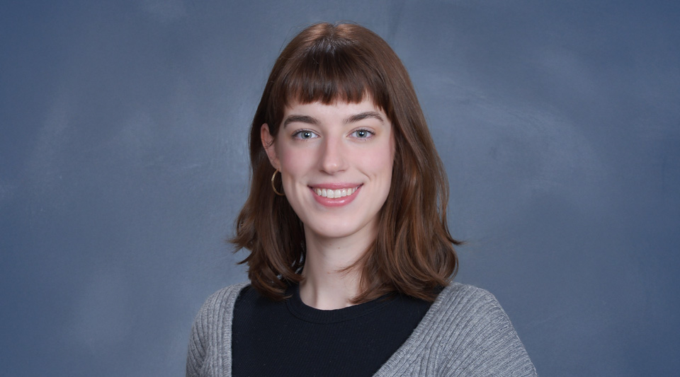 A young woman with auburn hair wearing a black shirt and gray sweater stands against a blue background and smiles