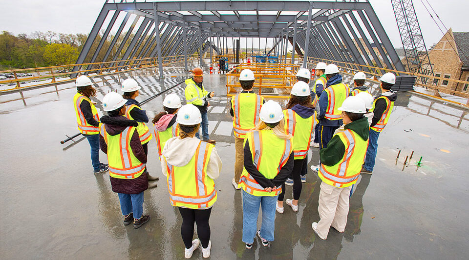 Students in hard hats and safety vests gather around an instructor at a construction site under a large steel frame.