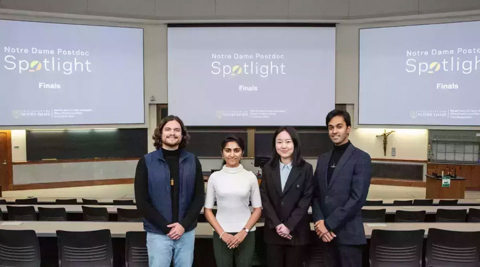 Four smiling Postdoc Spotlight finalists stand in a Notre Dame lecture hall. Screens behind them display "Notre Dame Postdoc Spotlight Finals" and university logos. A man in a blue vest, a woman in a white turtleneck, a woman in a black blazer, and a man in a dark suit are pictured.