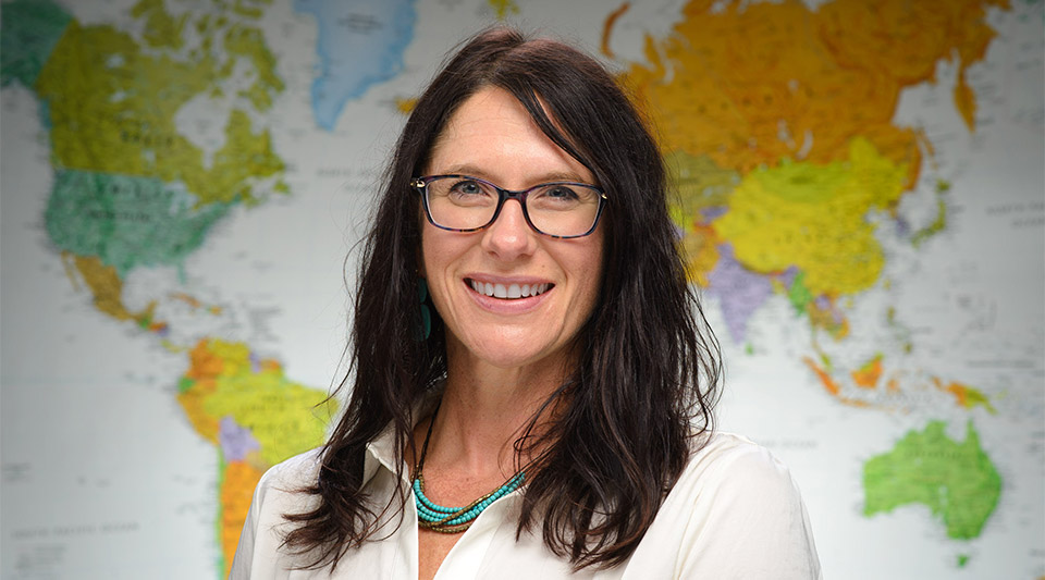 Tracy Kijewski-Correa, a woman with long, dark brown hair and glasses, stands in front of a colorful map.