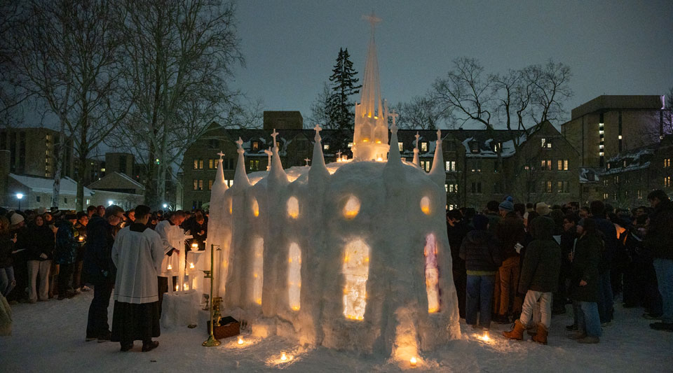 An image of the ice chapel at night with many people gathered for Mass and candles lighting the chapel.