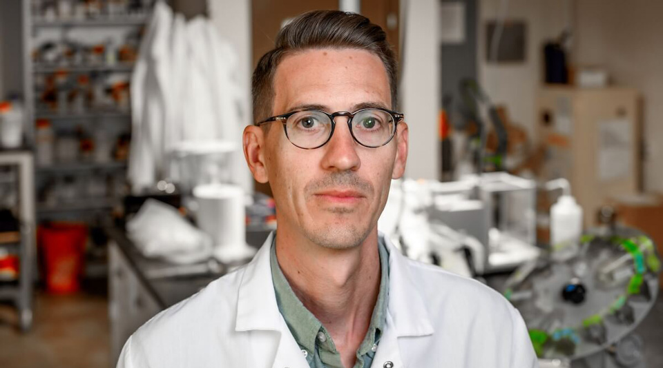 Kyle Doudrick wearing glasses and a white lab coat over a light green shirt, standing with a neutral expression in a laboratory.
