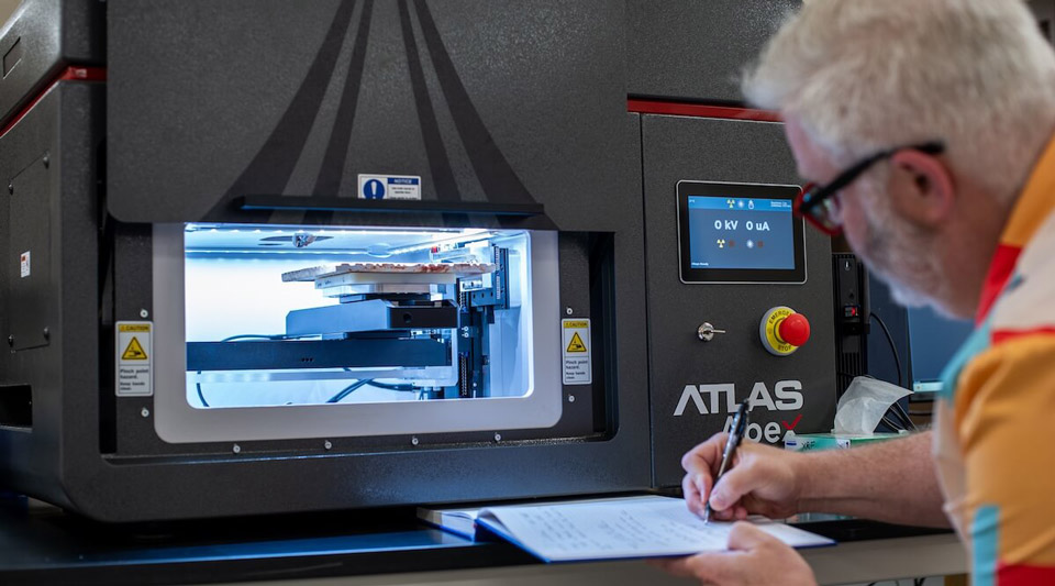 Gray-haired man in red glasses writes data, observing samples inside a blue-lit ATLAS X-ray machine.